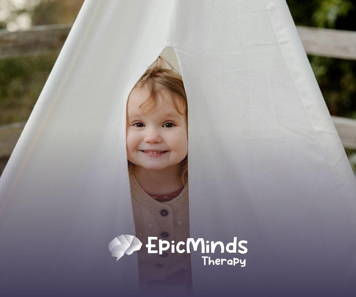 Smiling autistic toddler peeking out from inside a white play tent outdoors during ABA therapy in NC.