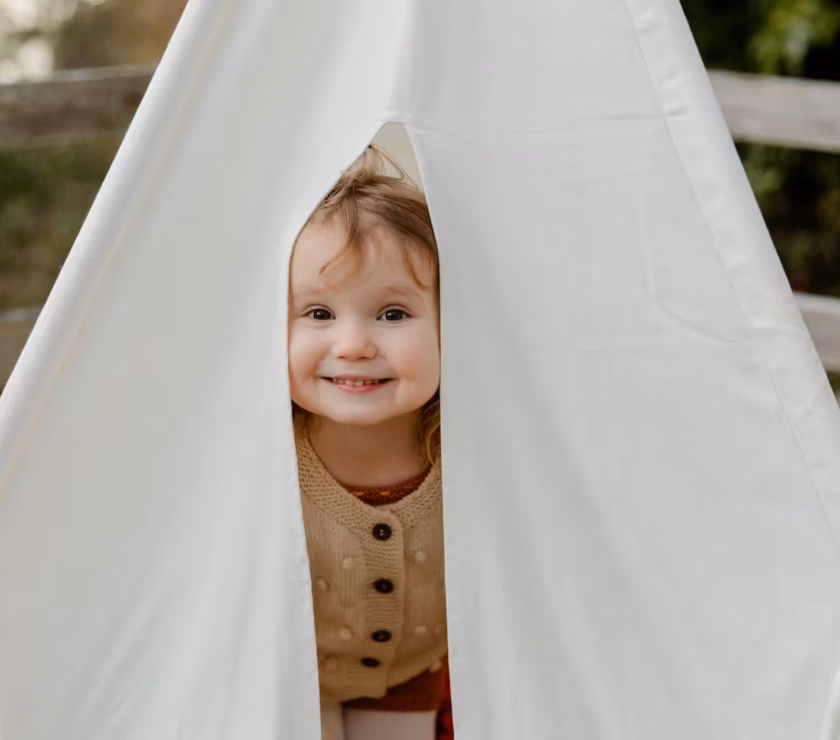 Smiling autistic toddler peeking out from inside a white play tent outdoors during ABA therapy in NC.