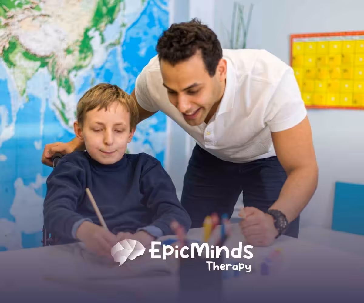 A smiling adult assists a child in a wheelchair with painting in a classroom setting. A world map and colorful calendar are visible in the background.
