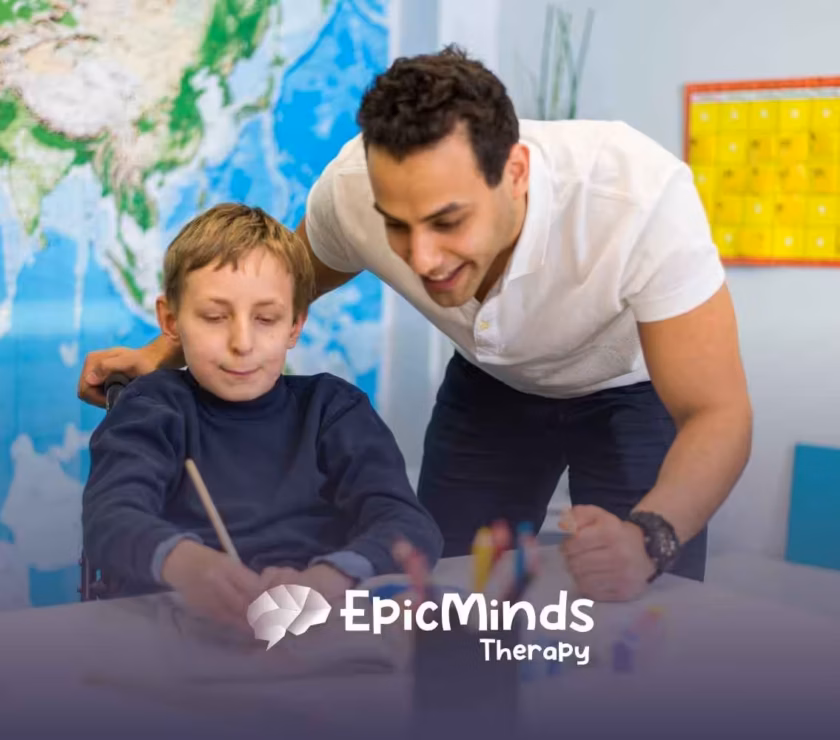 A smiling adult assists a child in a wheelchair with painting in a classroom setting. A world map and colorful calendar are visible in the background.