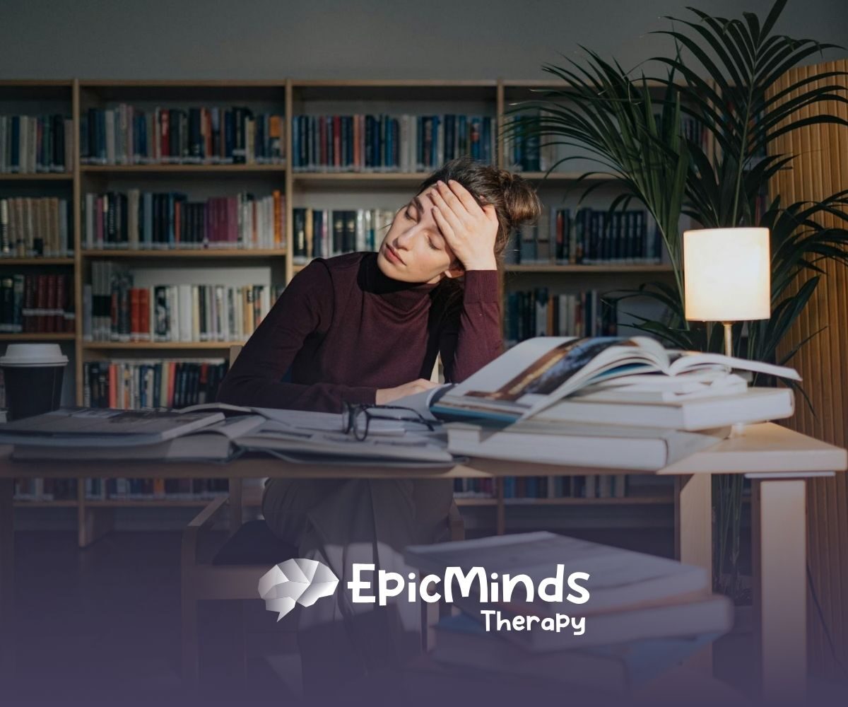 BCBA student in library looking tired while studying, surrounded by open books and a lamp in MD.
