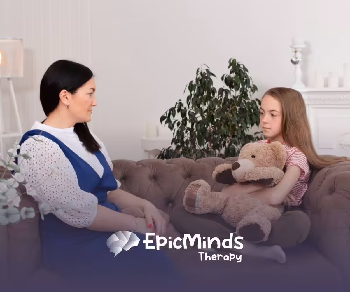 An autistic girl holding a teddy bear while talking to a BCBA on a couch during ABA therapy.