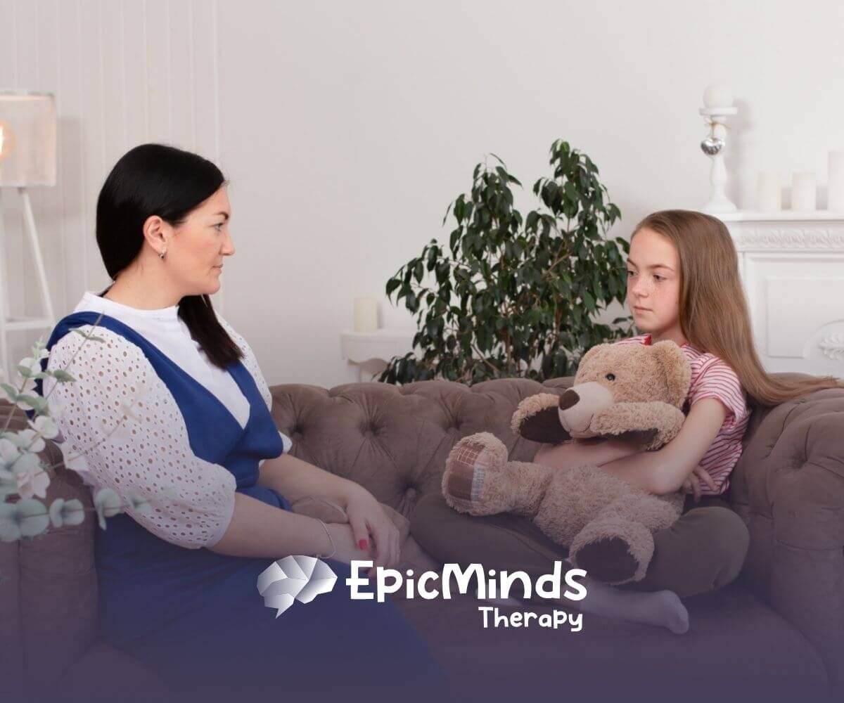 An autistic girl holding a teddy bear while talking to a BCBA on a couch during ABA therapy.