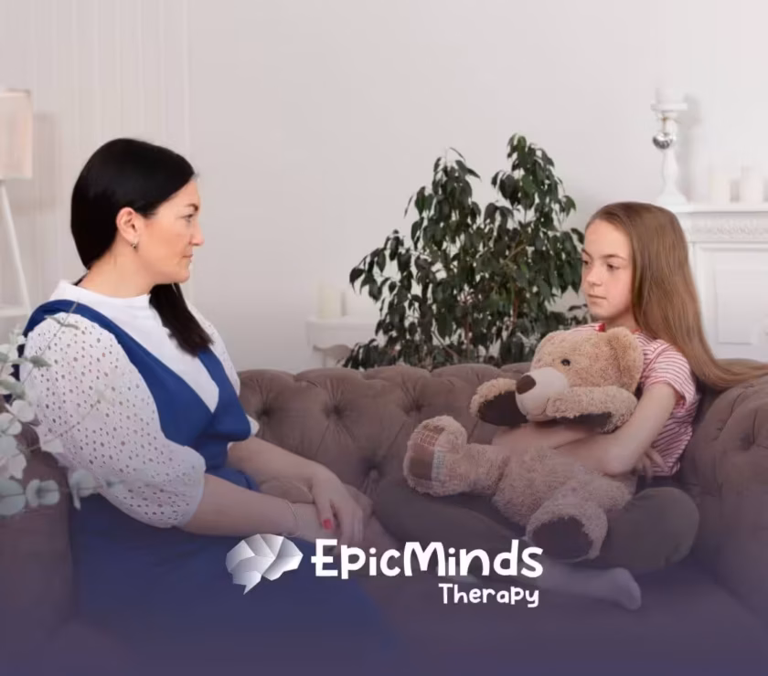 An autistic girl holding a teddy bear while talking to a BCBA on a couch during ABA therapy.