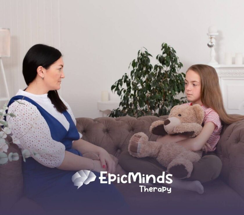 An autistic girl holding a teddy bear while talking to a BCBA on a couch during ABA therapy.