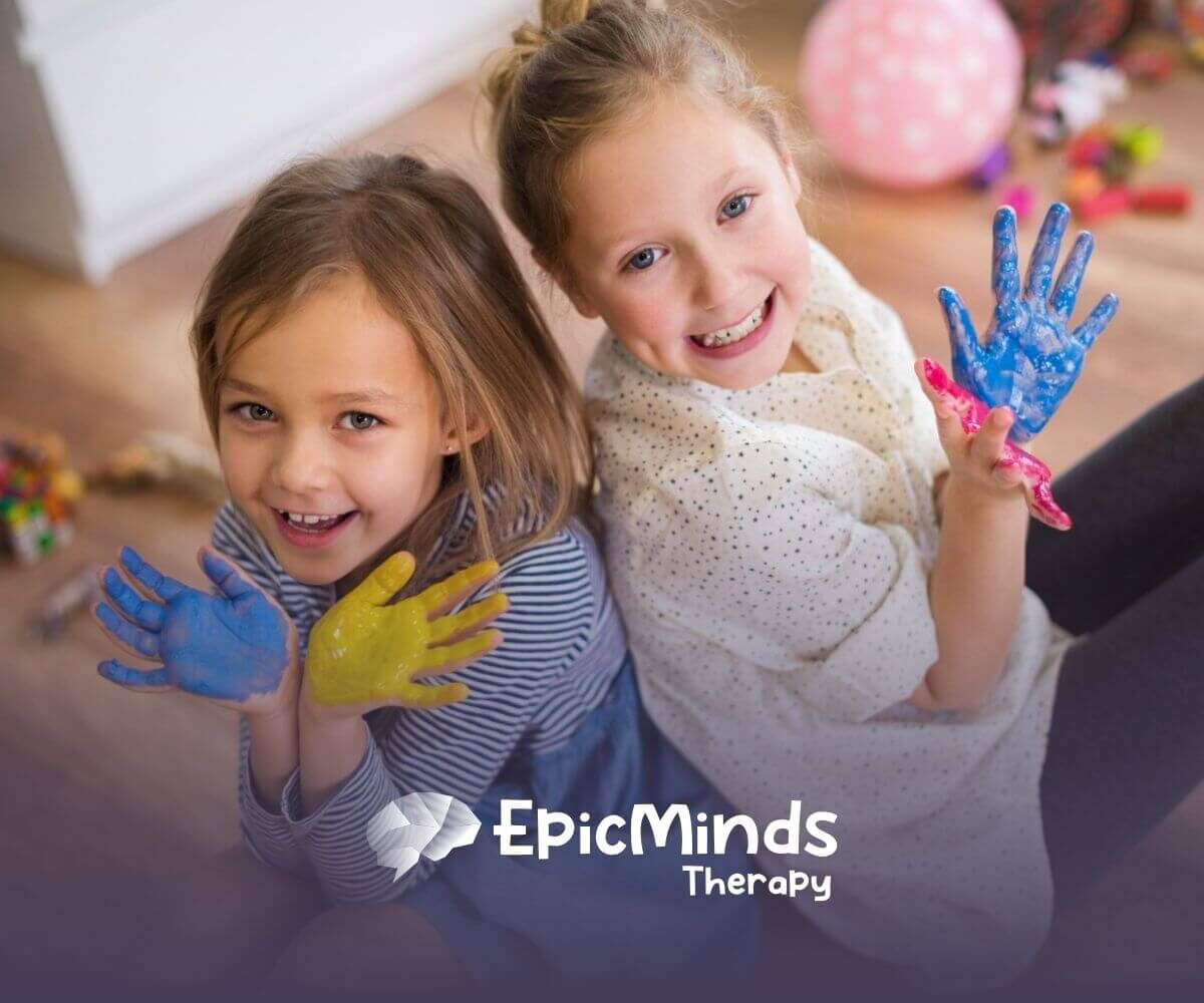 Two autistic girls sitting on the floor smiling and showing their hands covered in colorful paint during ABA therapy.
