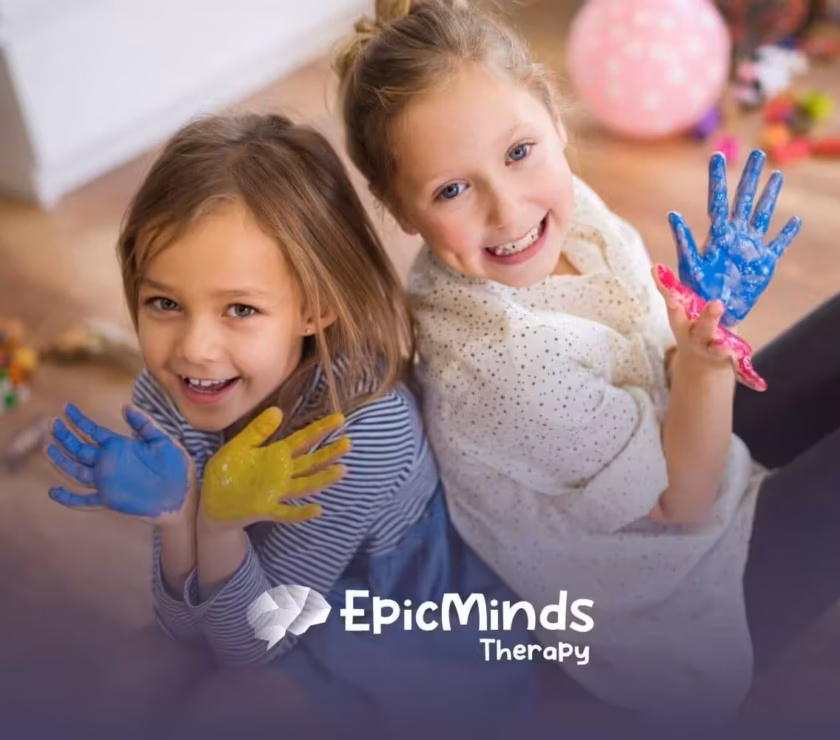 Two autistic girls sitting on the floor smiling and showing their hands covered in colorful paint during ABA therapy.