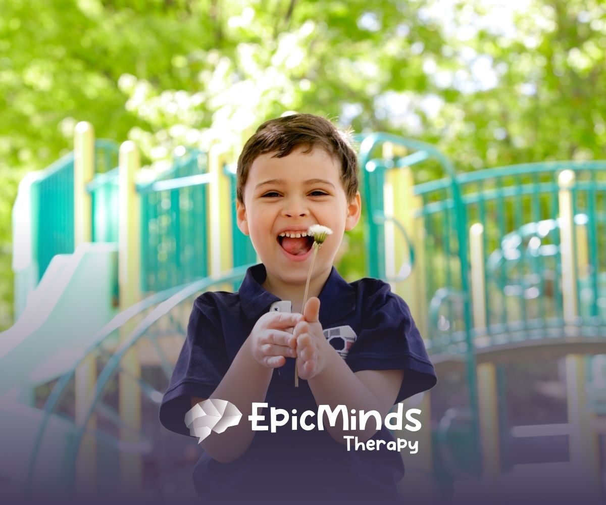 A joyful child holds a white flower, smiling widely against a vibrant playground backdrop. Sunlight filters through green leaves, creating a cheerful atmosphere.