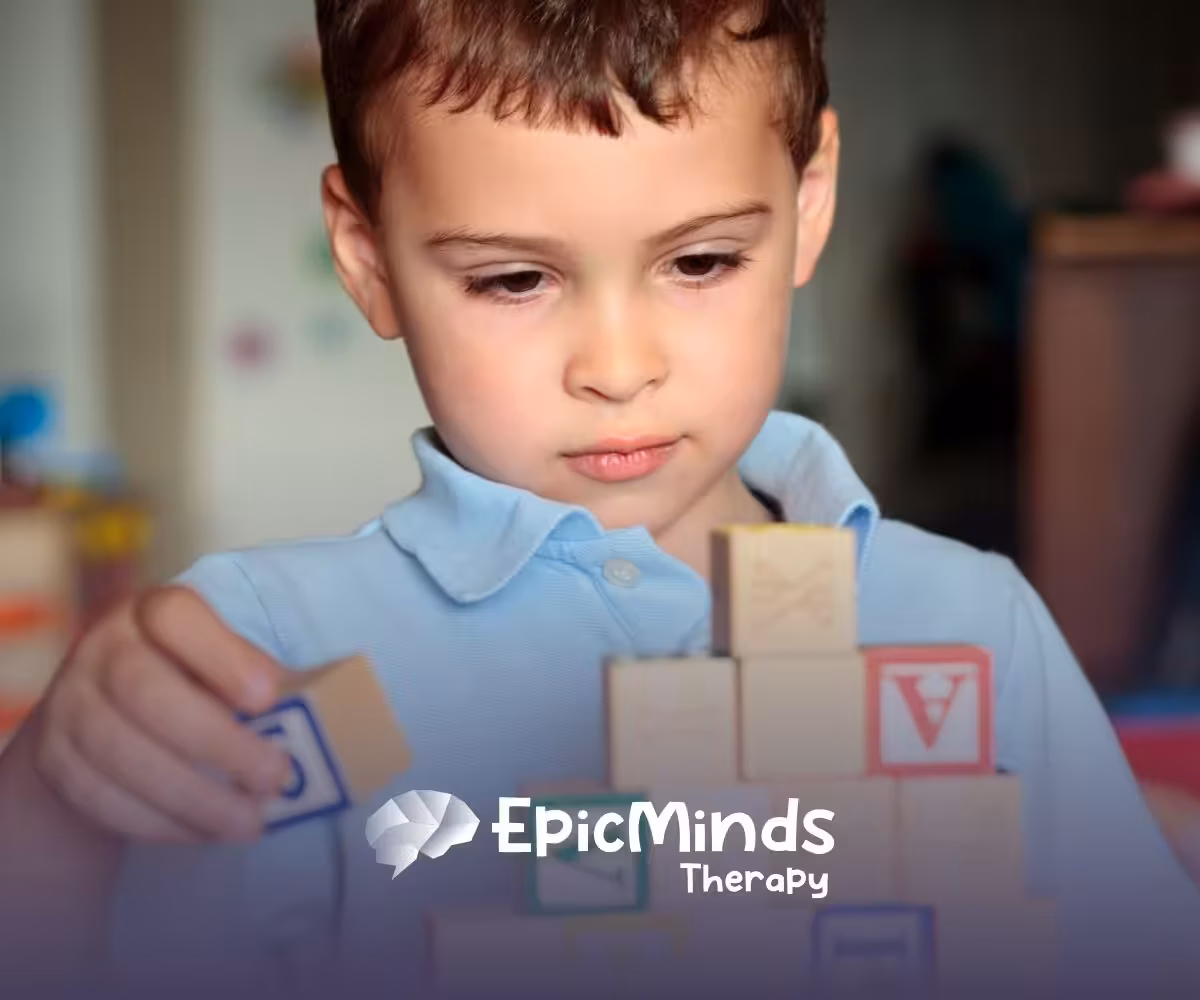 A young child in a blue shirt focuses intently on stacking colorful wooden blocks. The scene conveys concentration and creativity in a playful setting.