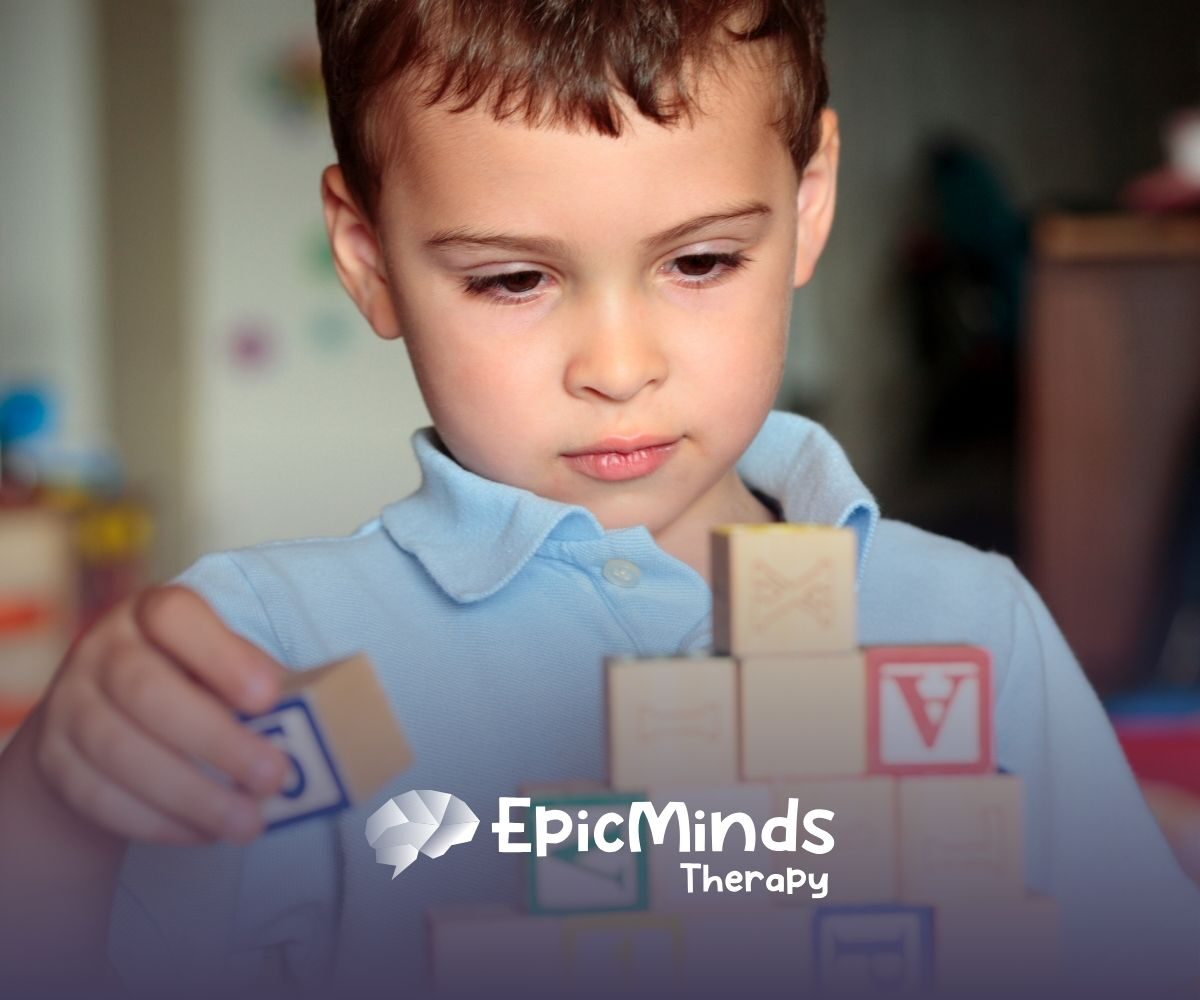 A young child in a blue shirt focuses intently on stacking colorful wooden blocks. The scene conveys concentration and creativity in a playful setting.