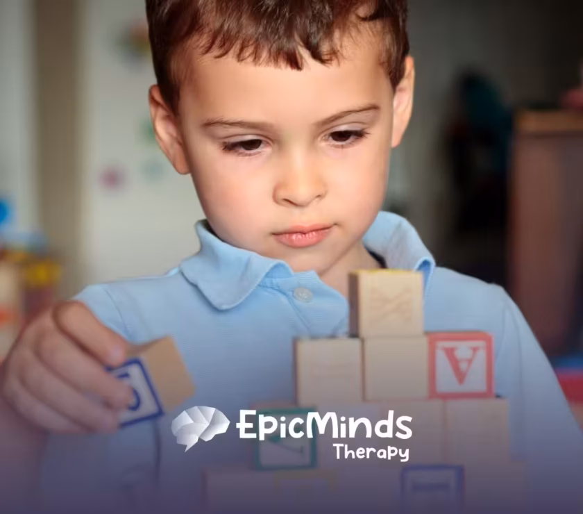 A young child in a blue shirt focuses intently on stacking colorful wooden blocks. The scene conveys concentration and creativity in a playful setting.