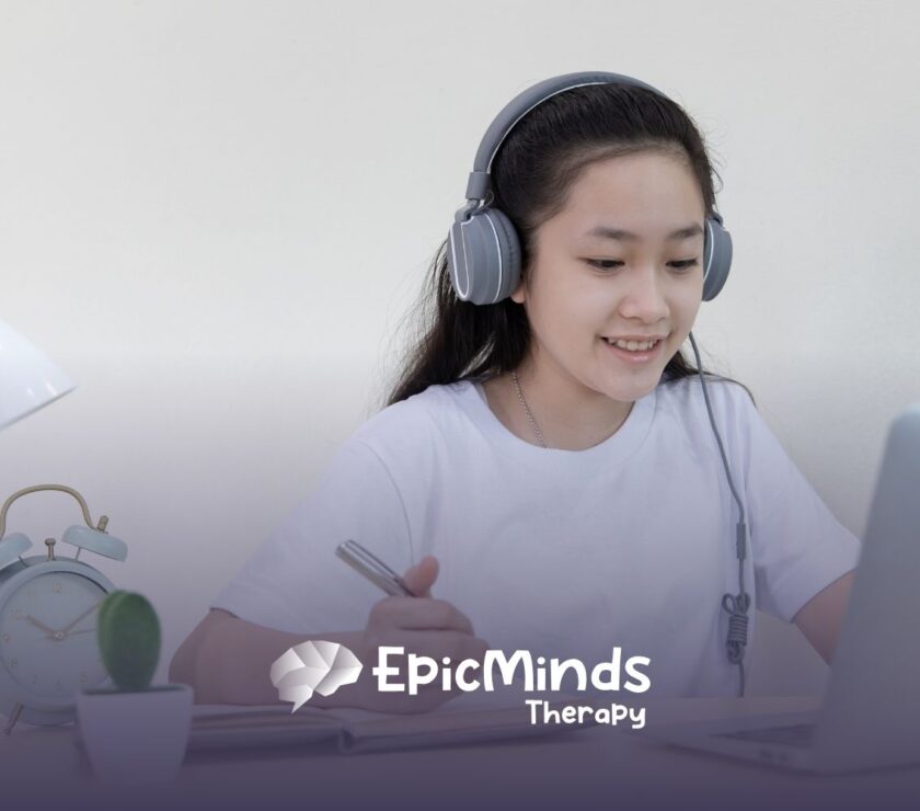A smiling girl wearing headphones studies at a laptop, writing in a notebook. A desk lamp, alarm clock, and small cactus are beside her, conveying focus.