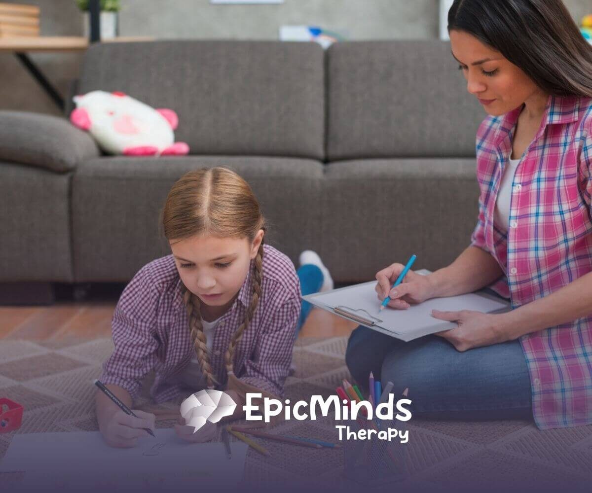 Autistic girl drawing on paper on the floor while ABA therapist beside her takes notes during ABA therapy.
