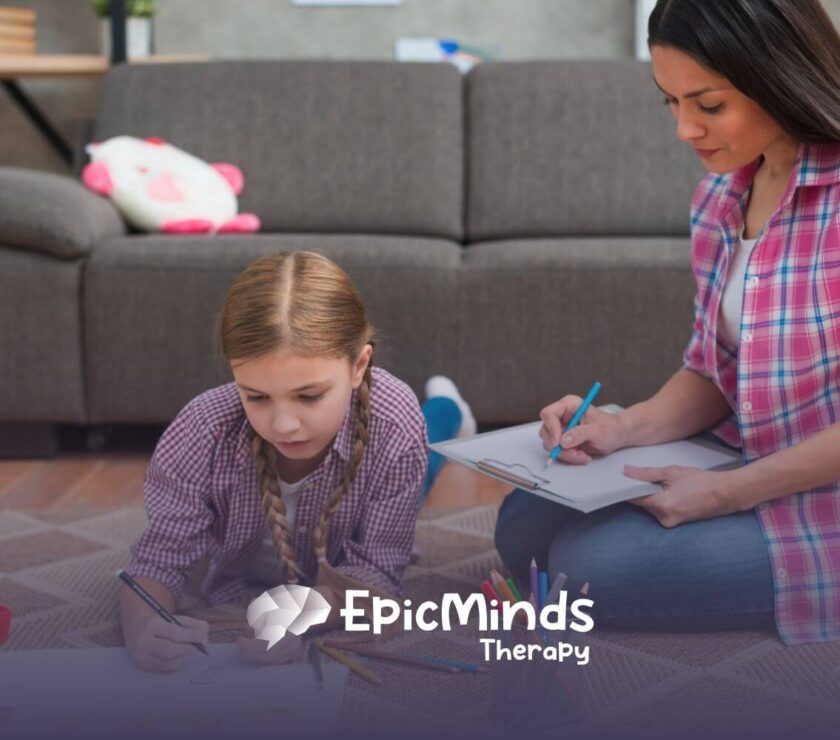 Autistic girl drawing on paper on the floor while ABA therapist beside her takes notes during ABA therapy.