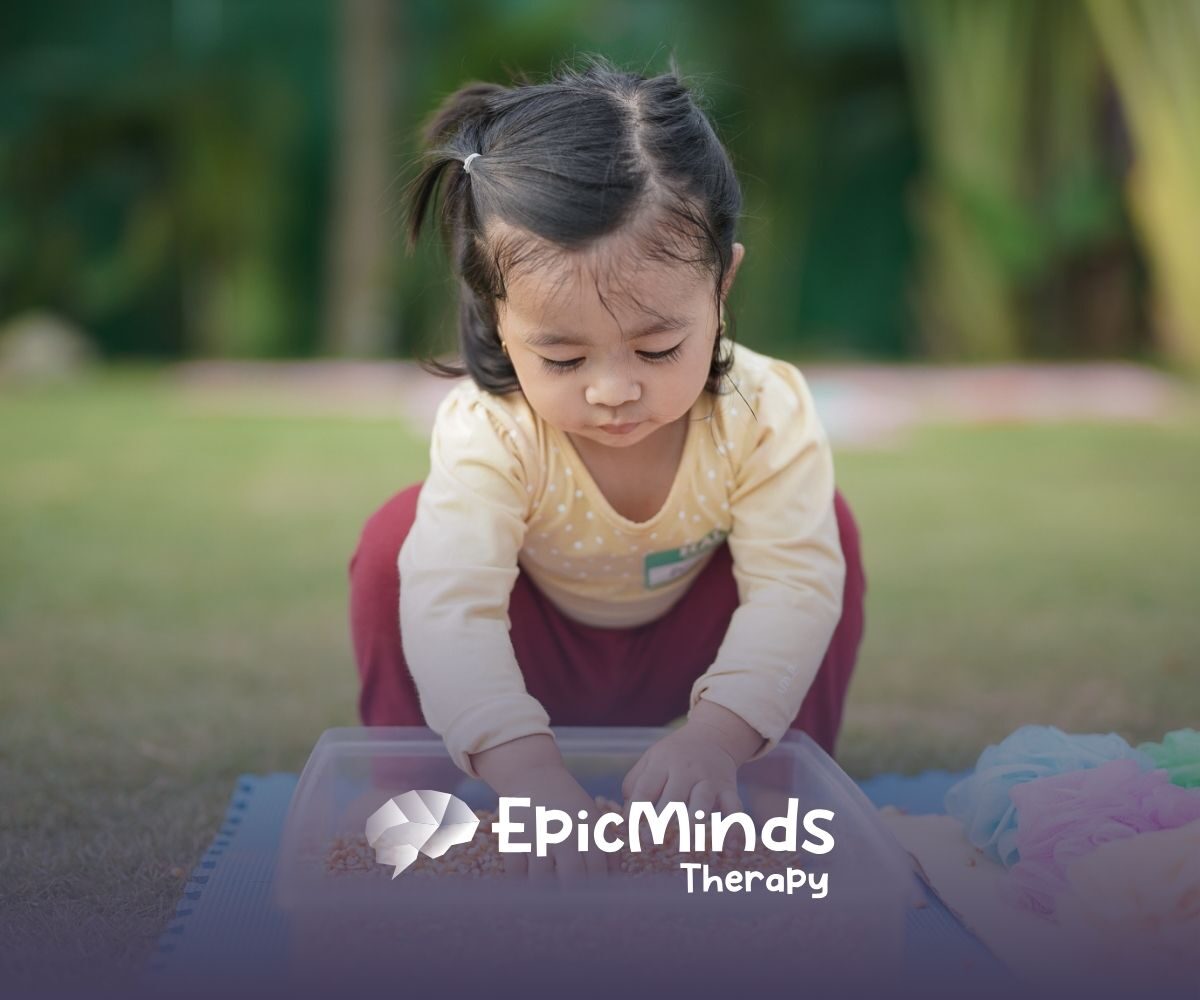 A young girl with pigtails intently plays with grains in a plastic bin outdoors. She wears a yellow shirt and red pants, exuding curiosity and focus.