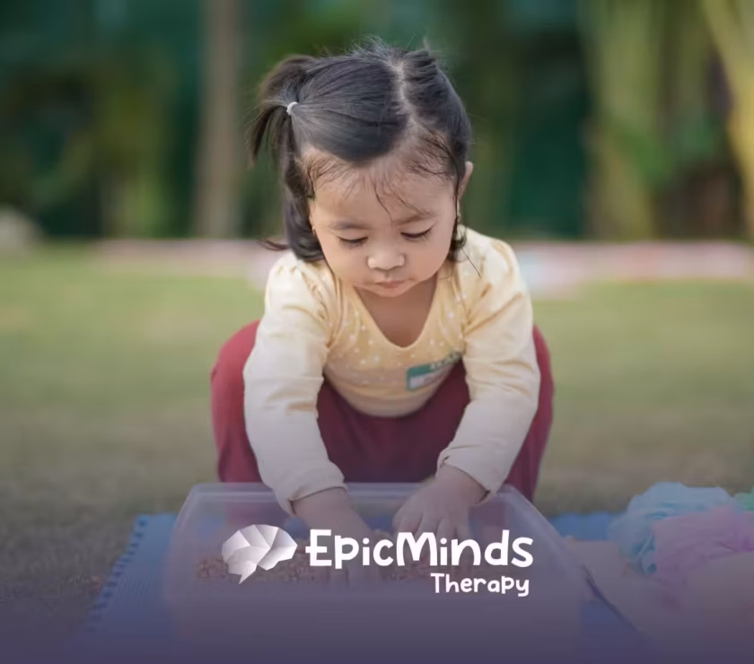 A young girl with pigtails intently plays with grains in a plastic bin outdoors. She wears a yellow shirt and red pants, exuding curiosity and focus.