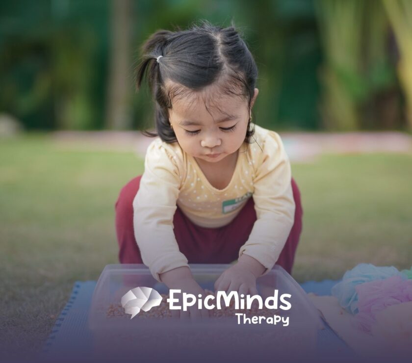 A young girl with pigtails intently plays with grains in a plastic bin outdoors. She wears a yellow shirt and red pants, exuding curiosity and focus.