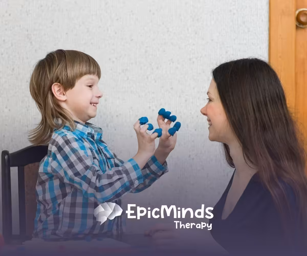 A smiling boy shows his fingers covered in blue modeling clay to an ABA therapist in ABA therapy.