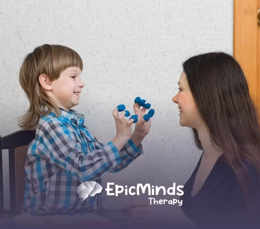 A smiling boy shows his fingers covered in blue modeling clay to an ABA therapist in ABA therapy.