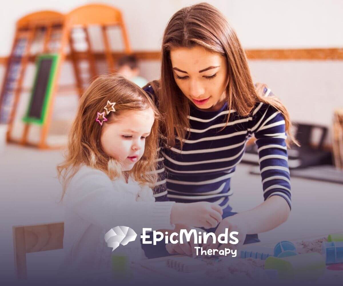 An ABA therapist helps a little girl play with kinetic sand and toys at a table in ABA therapy.
