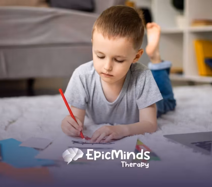 A boy with autism lies on the floor, drawing with a red pencil in ABA therapy.