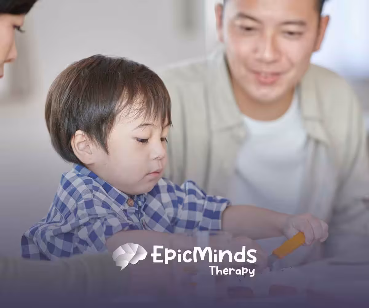 A young child in a blue plaid shirt focuses intently on a meal, using an orange utensil. Two smiling adults are nearby.