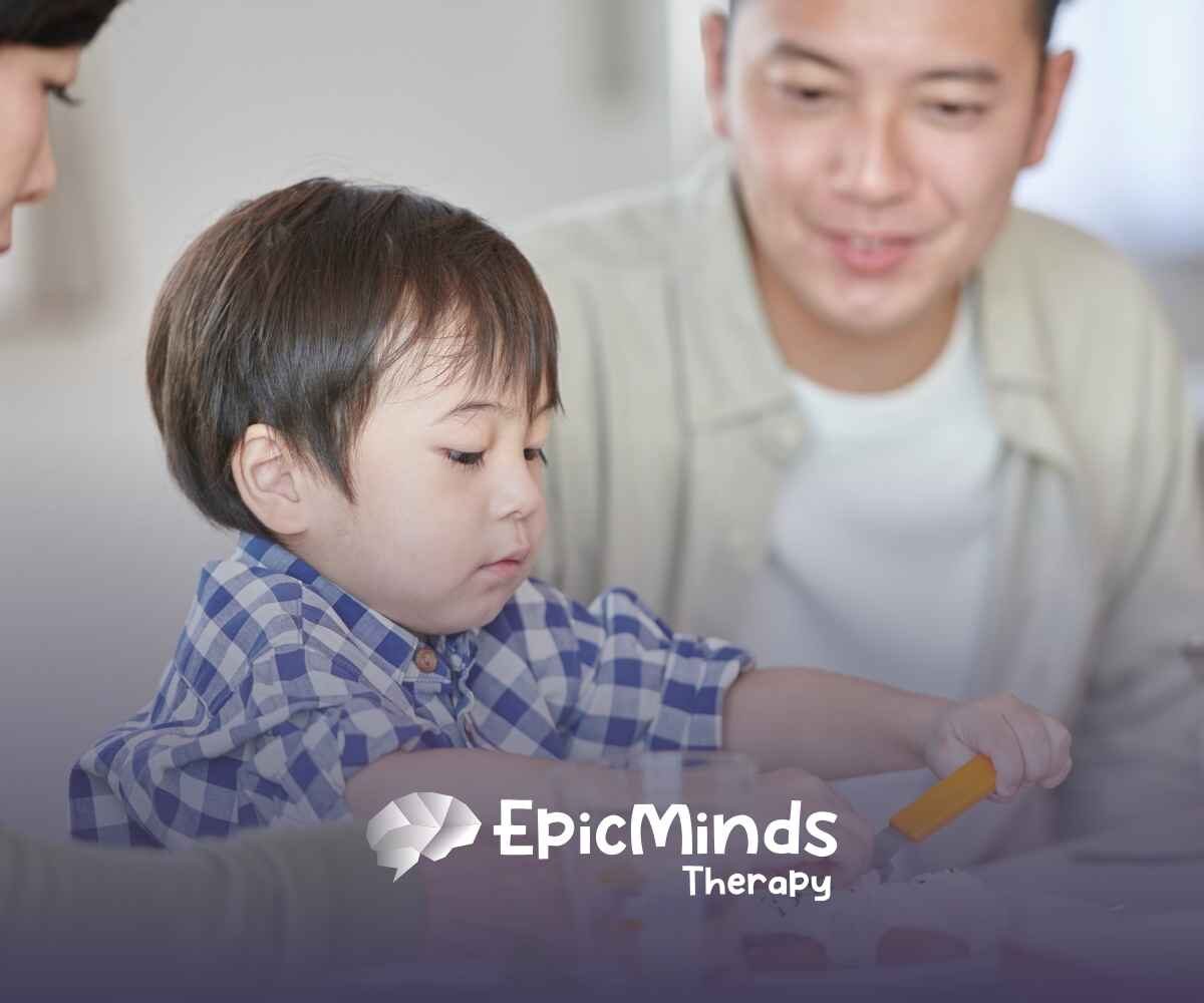 A young child in a blue plaid shirt focuses intently on a meal, using an orange utensil. Two smiling adults are nearby.