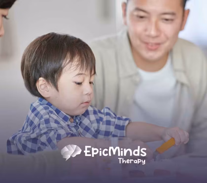 A young child in a blue plaid shirt focuses intently on a meal, using an orange utensil. Two smiling adults are nearby.
