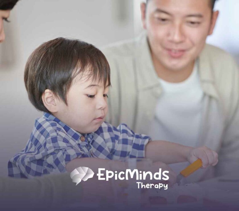A young child in a blue plaid shirt focuses intently on a meal, using an orange utensil. Two smiling adults are nearby.