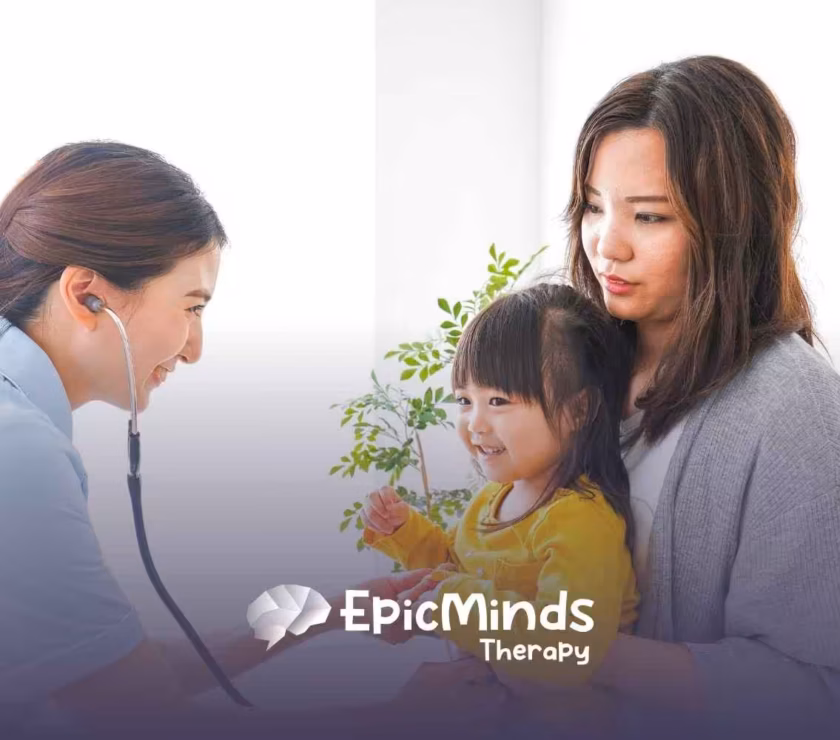 A nurse with a stethoscope smiles at a smiling toddler in a yellow shirt, held by a woman. The setting is bright and caring, with a small plant in the background.