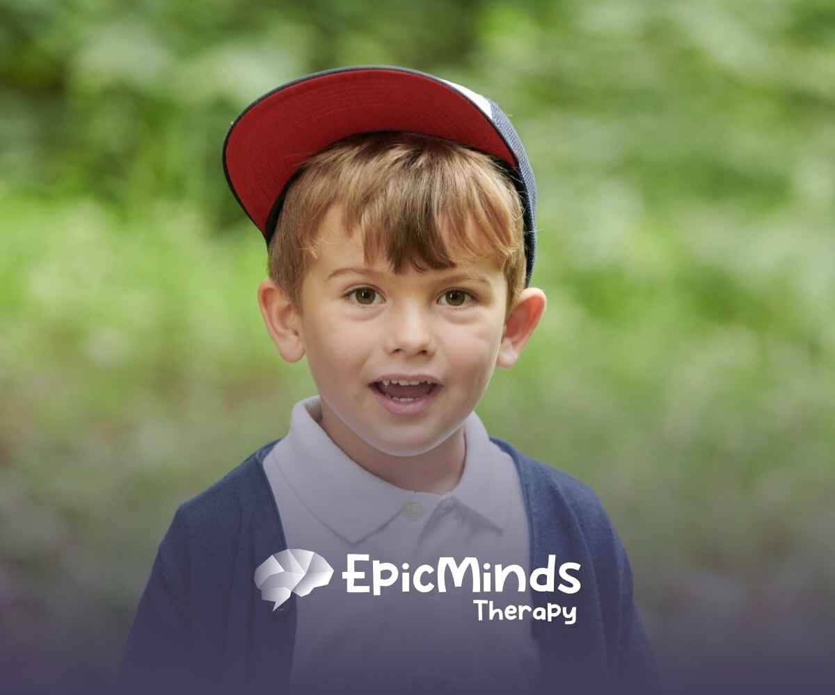Young boy in a red cap, white shirt, and navy cardigan smiles in a green outdoor setting.