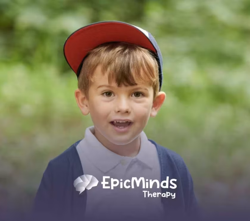 Young boy in a red cap, white shirt, and navy cardigan smiles in a green outdoor setting.