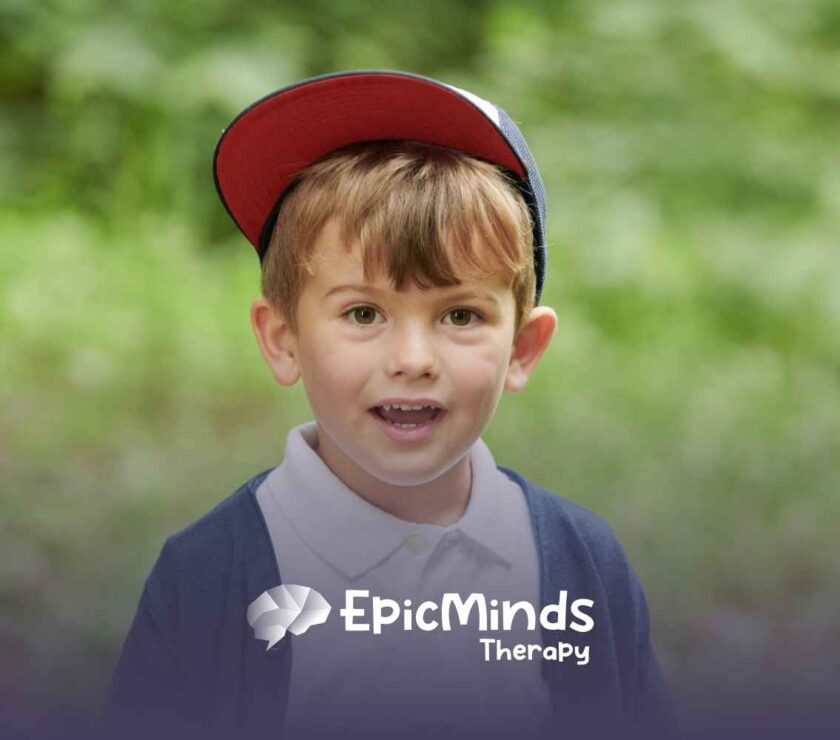 Young boy in a red cap, white shirt, and navy cardigan smiles in a green outdoor setting.