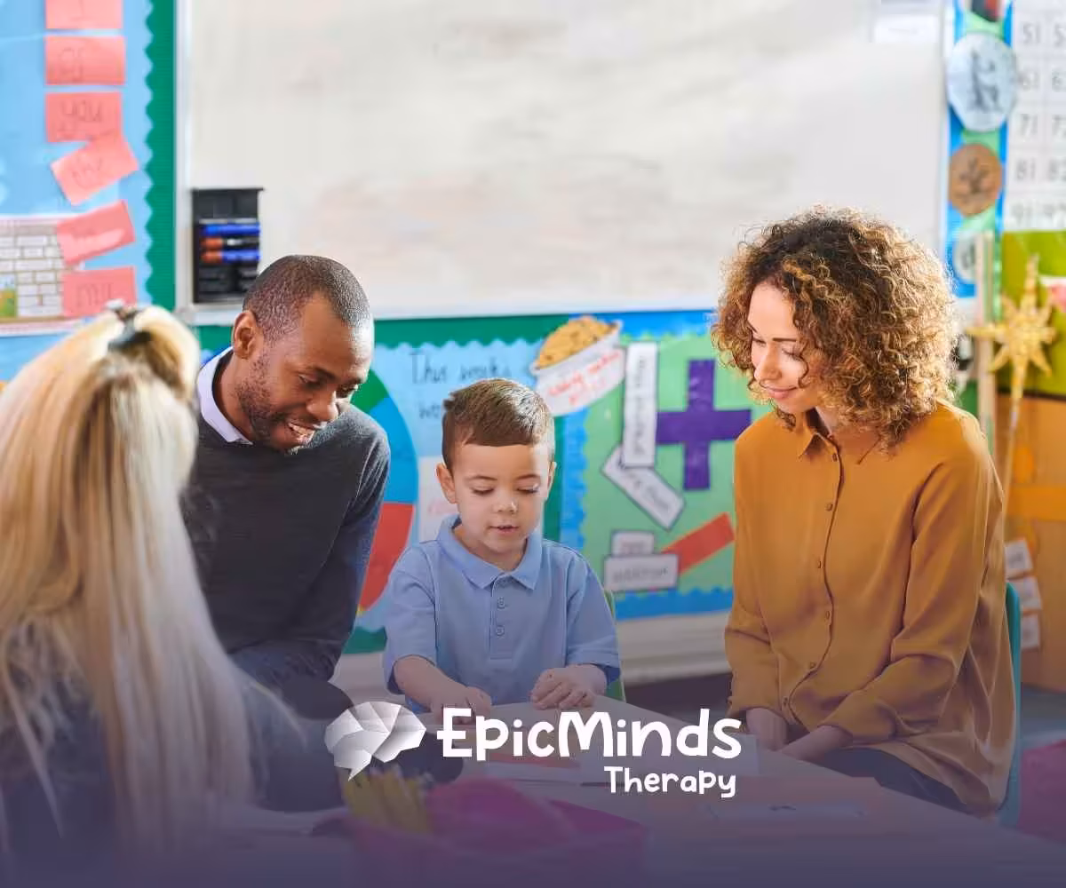 A young boy in a blue shirt is engaged with three adults in a colorful classroom setting. The mood is warm and supportive, with educational posters in the background.
