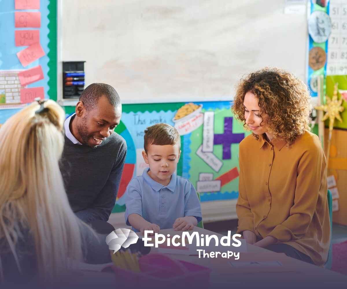 A young boy in a blue shirt is engaged with three adults in a colorful classroom setting. The mood is warm and supportive, with educational posters in the background.