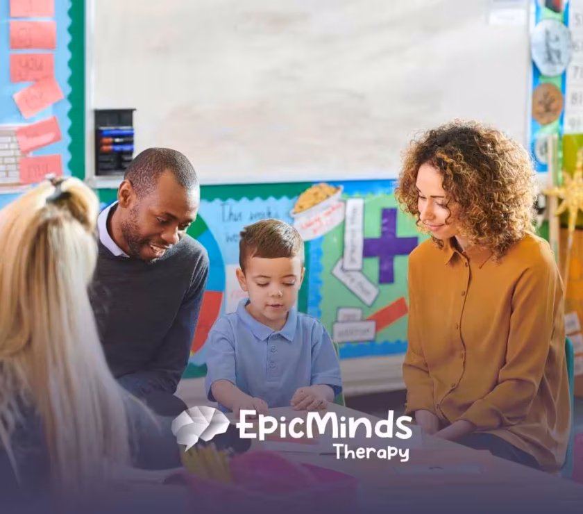 A young boy in a blue shirt is engaged with three adults in a colorful classroom setting. The mood is warm and supportive, with educational posters in the background.