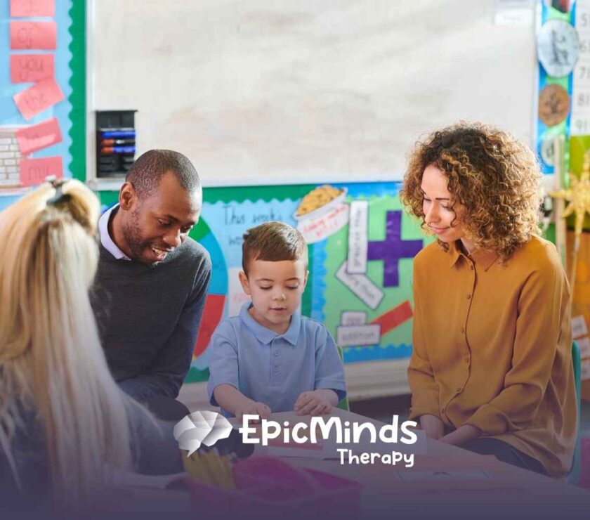 A young boy in a blue shirt is engaged with three adults in a colorful classroom setting. The mood is warm and supportive, with educational posters in the background.