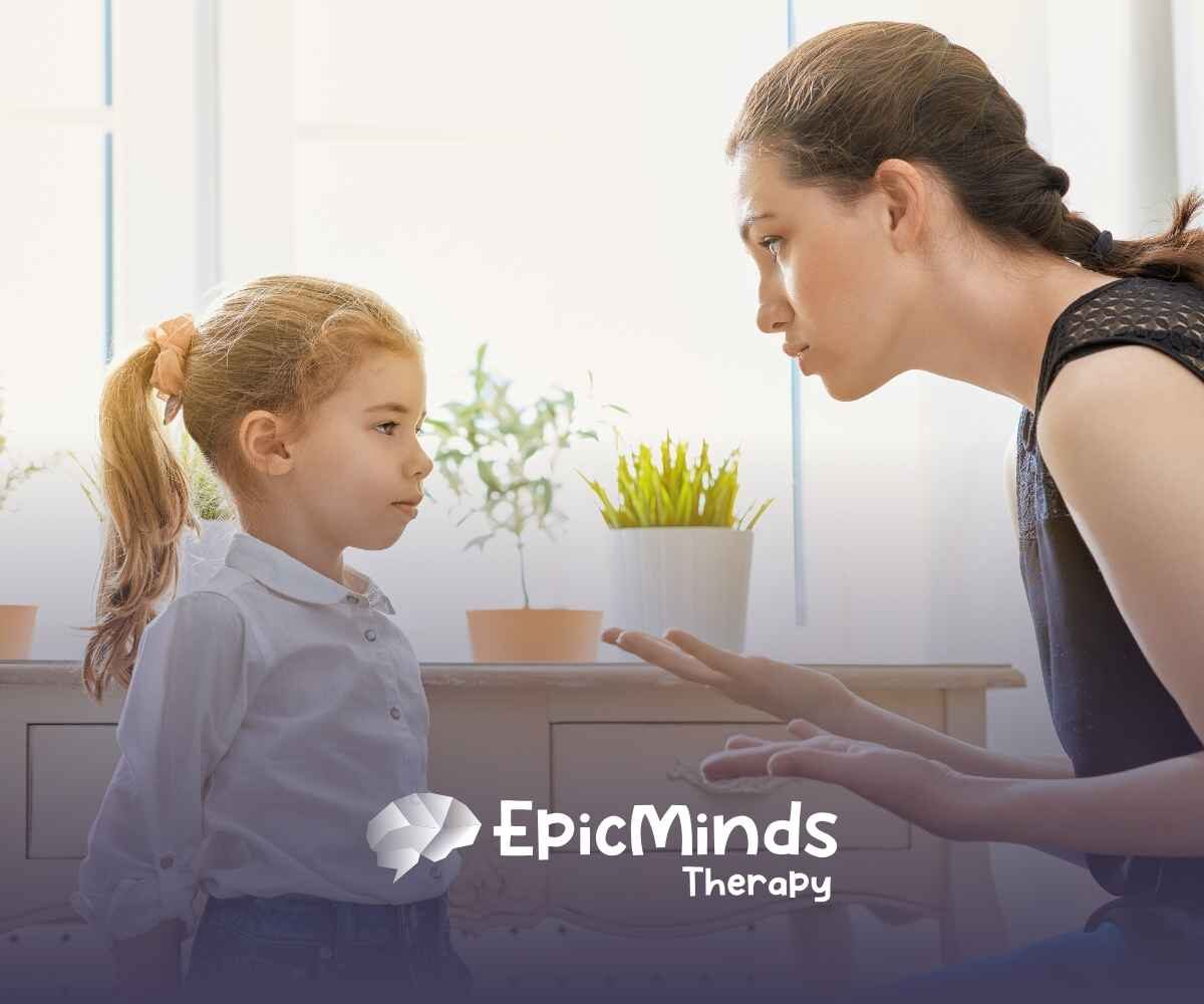 A woman kneels beside a young girl, speaking gently. The girl listens intently. Potted plants sit on a table in the bright, softly lit room.