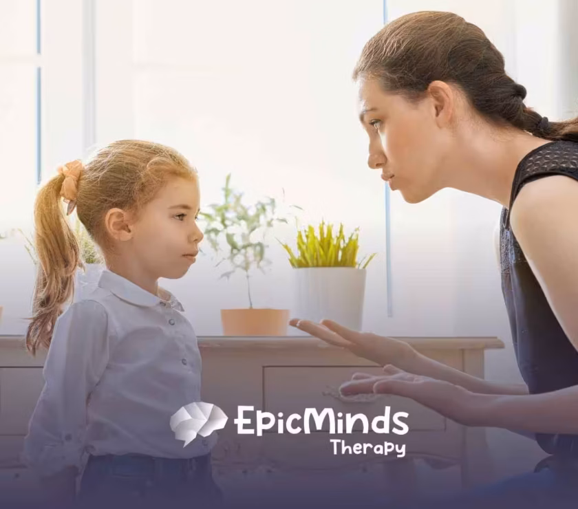 A woman kneels beside a young girl, speaking gently. The girl listens intently. Potted plants sit on a table in the bright, softly lit room.