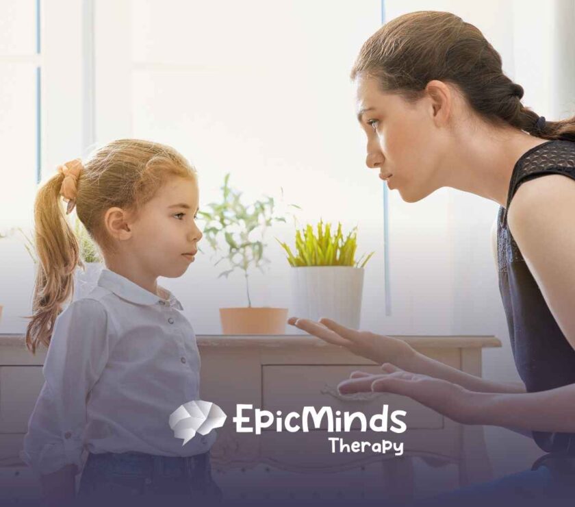 A woman kneels beside a young girl, speaking gently. The girl listens intently. Potted plants sit on a table in the bright, softly lit room.