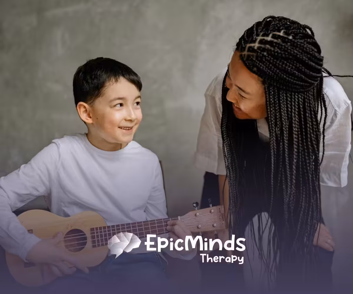 An autistic boy smiling while playing a small guitar as an RBT with braids leans toward him during ABA therapy in NC.