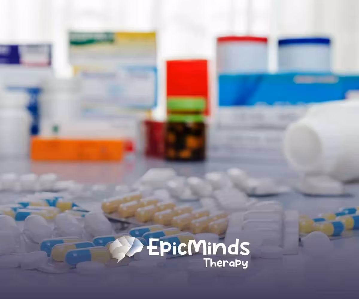 A variety of pills and capsules are scattered on a white surface, with medicine boxes blurred in the background. The image conveys a clinical or pharmaceutical setting.