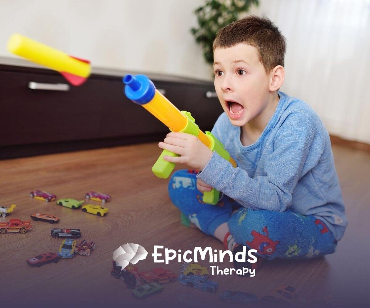 Young boy with autism sitting on the floor excitedly playing with a toy blaster and toy cars.