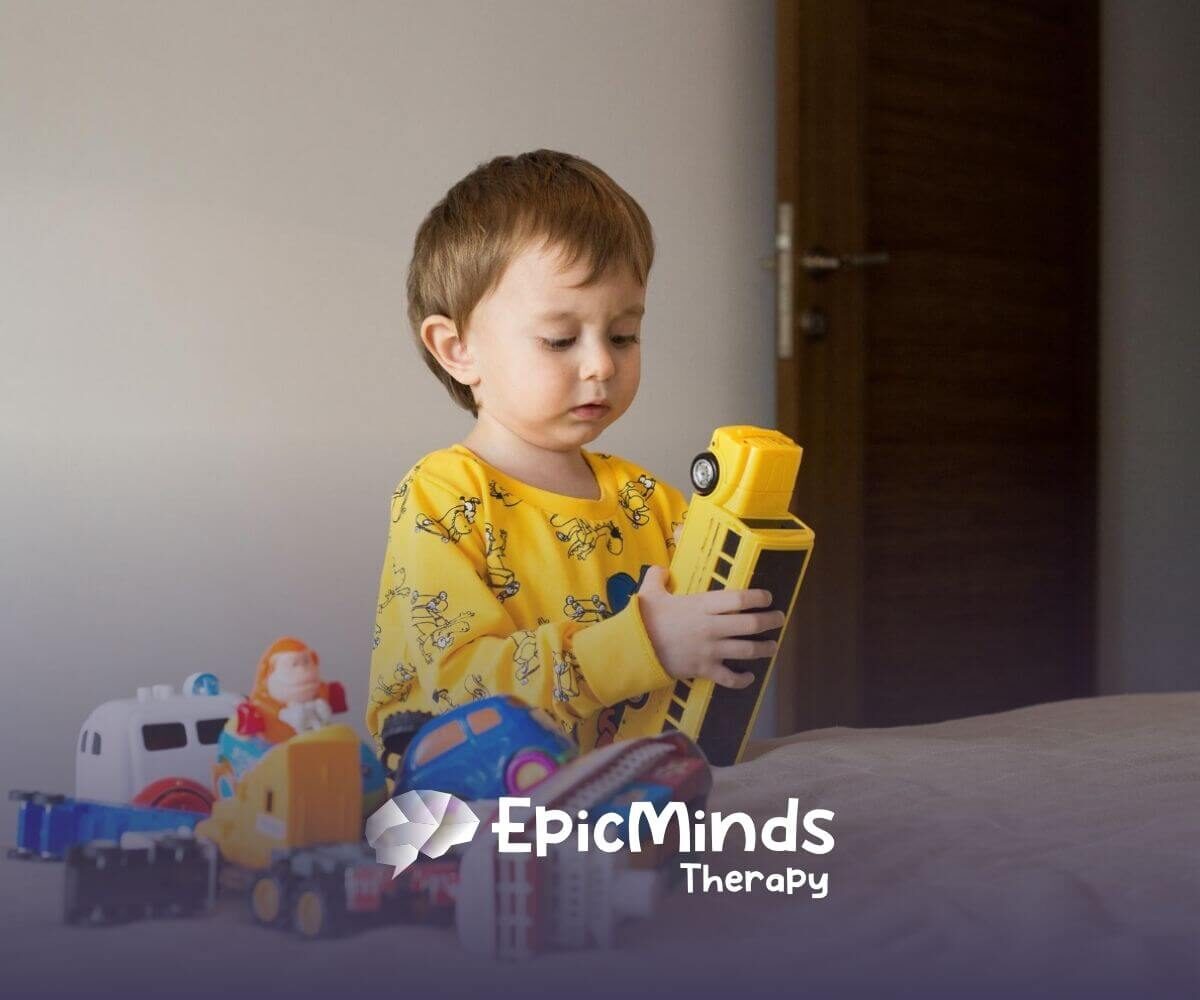 Toddler with autism holding a yellow toy bus while playing with toy vehicles.