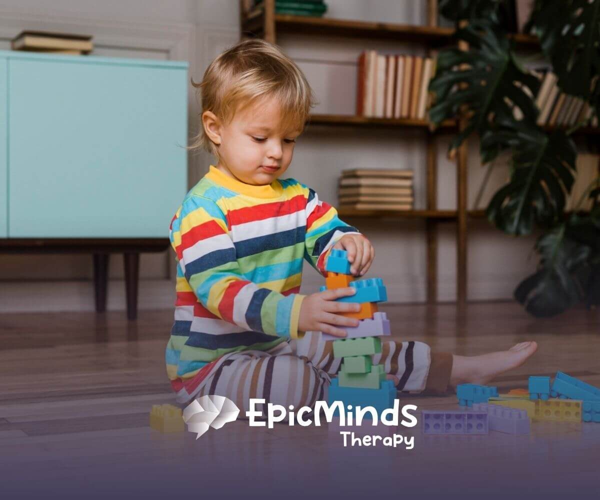 A toddler with autism stacking bright plastic blocks on the floor at home.