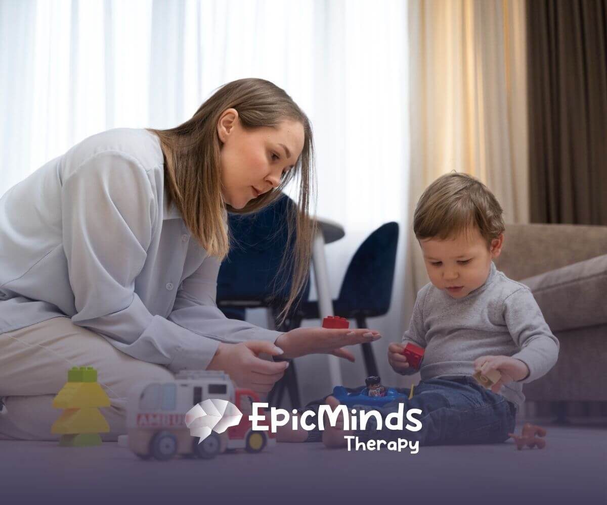 A BCBA sitting on the floor helping a toddler with autism play with toy blocks.