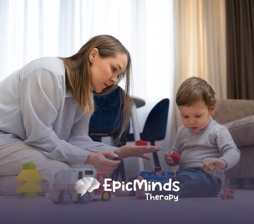 A BCBA sitting on the floor helping a toddler with autism play with toy blocks.
