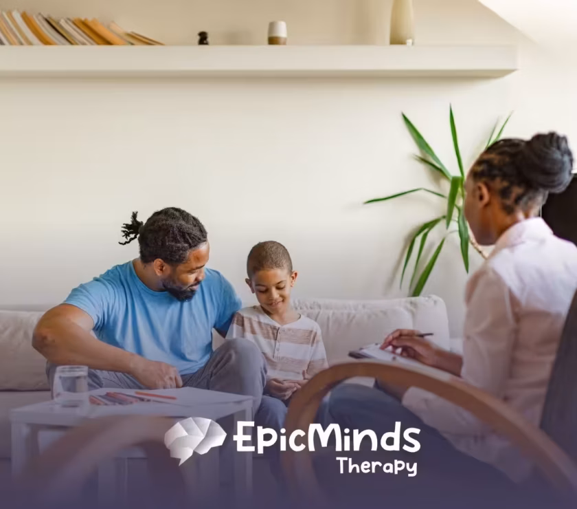 A therapist sits with a father and son on a couch in a calm, well-lit room. The boy looks at a notebook while the father and therapist engage with him.