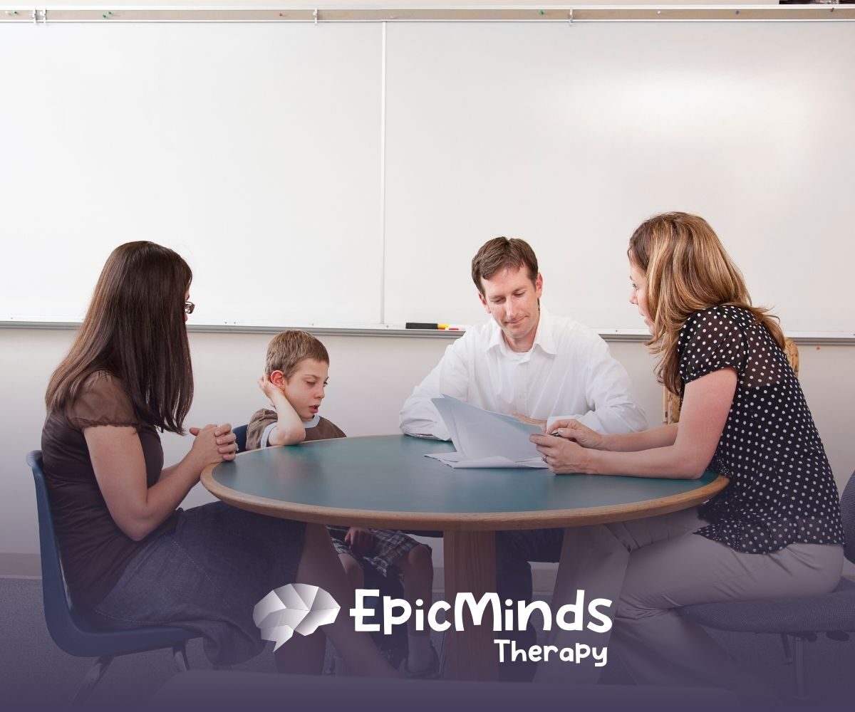 A woman and a man sit at a round table with a young boy and his teacher, reviewing documents in a room with a white board.