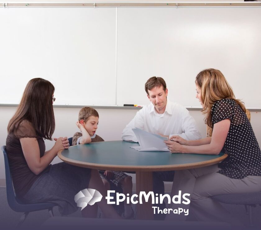 A woman and a man sit at a round table with a young boy and his teacher, reviewing documents in a room with a white board.
